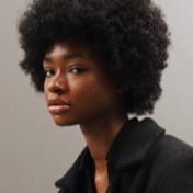 Mulher com cabelos crespos em estilo black power, vestindo camisa preta, posando para uma foto de perfil profissional.